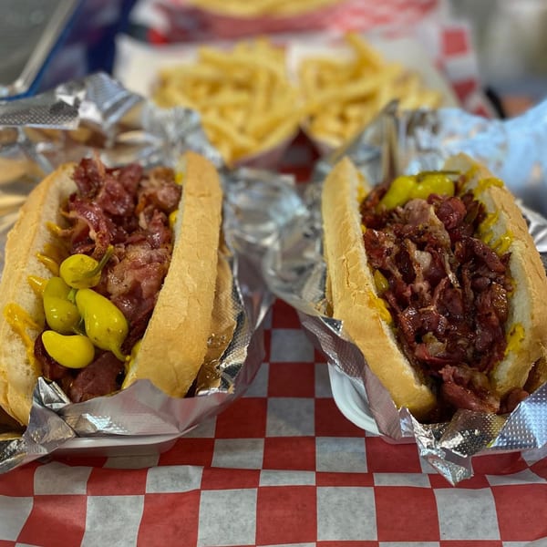 Two hot dogs with pastrami, mustard, and peppers, with fries in the background.