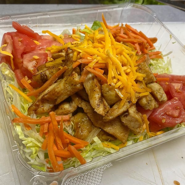 Grilled chicken salad with shredded lettuce, tomatoes, carrots, and cheddar cheese in a plastic container.